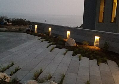 Modern house with outdoor lighting along a paved path leading to a coastal view at dusk.